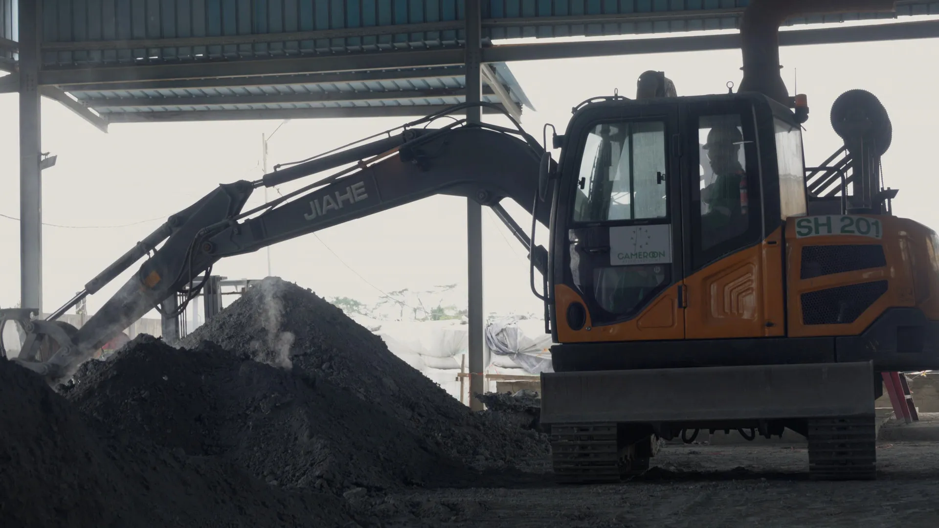 Excavator under a shed, in front of a heap of aluminium dross giving off steam