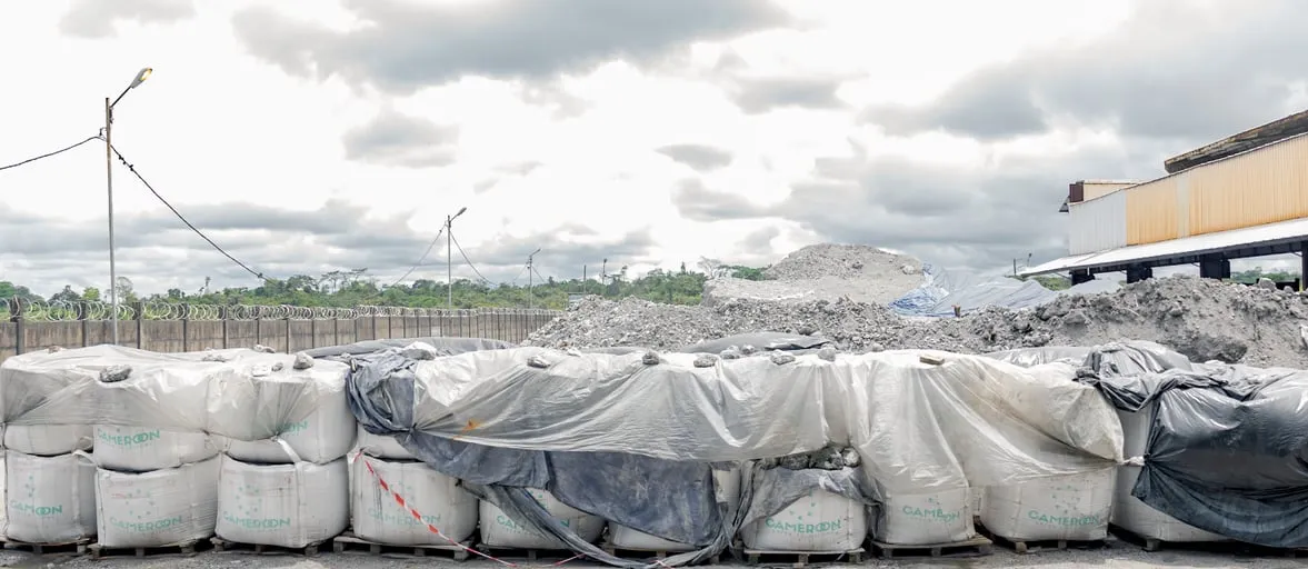 Row of large covered bags on pallets, in front of heaps of material, under a cloudy sky